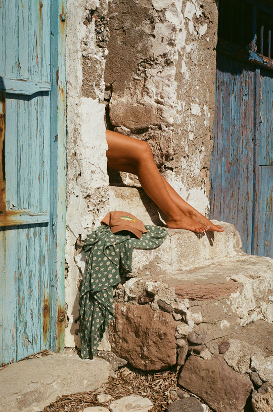 tanned legs, hat and sarong beside stone steps next to a blue timber door in Klima on the island of Milos. 