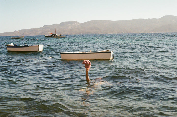 Man holding pink hat with fish embroidered on it our of the Adriatic Sea with little timber row boats bobbing in the distance. He is swimming off Klima, a little seaside town on the island of Milos, Greece