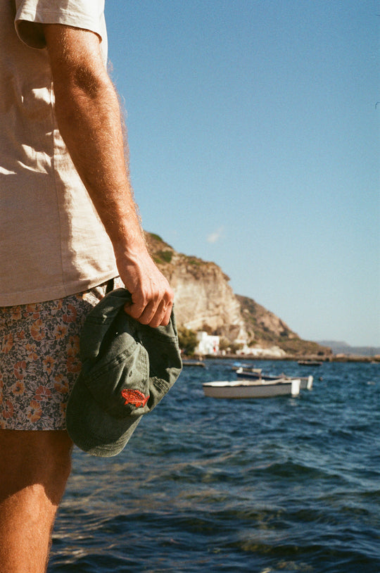 Man holds green hat with red coral trout fish embroidered on it. He is standing by the sea and little timber boats are seen in the distance. Location is the seaside town of Klima in Milos, Greece