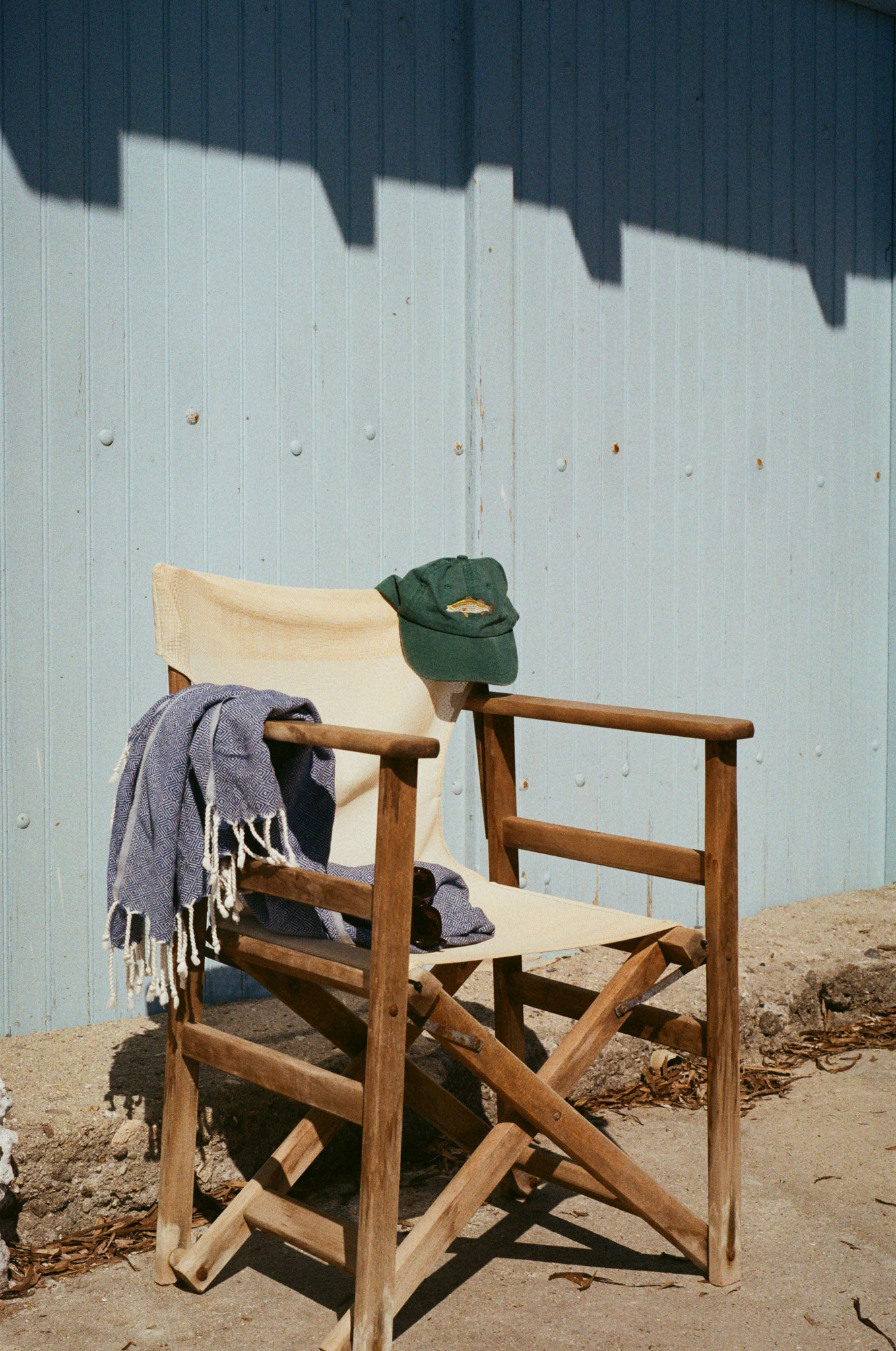 green hat with a barramundi fish embroidered on it is hung over the corner of a white directors chair against a blue timber boat door in Greece