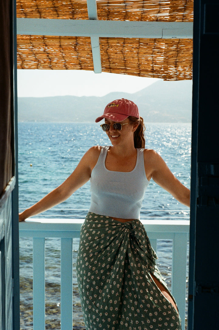 Ashleigh Bridget leans against a blue balcony overlooking the Adriatic Sea on the island of Milos. She is wearing a pink fish lid from Pallion Point, blue singlet and green patterned sarong. Location: Klima, Milos, Greece