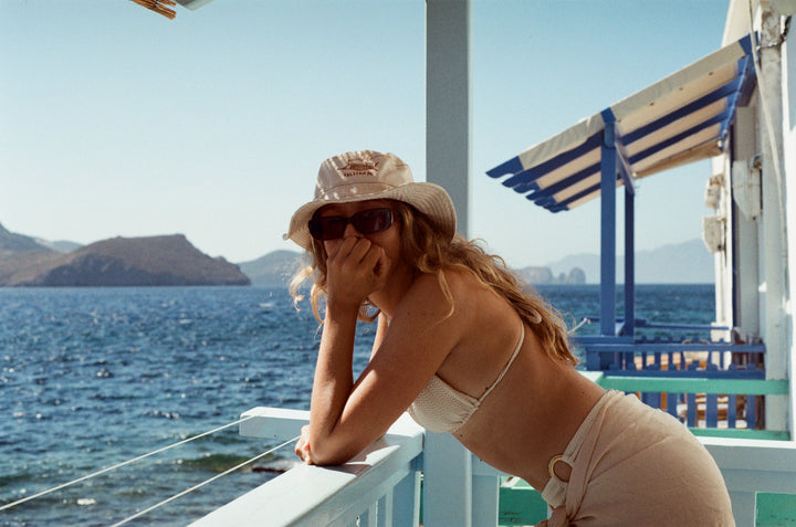 Female is leaning on a blue balcony looking at the camera. She is wearing a cream wide brim hat with a sailfish embroidered on it, sungllasses, cream bikinis and a sarong. In the background is the Adriatic Sea and the island of Milos. Location Klima